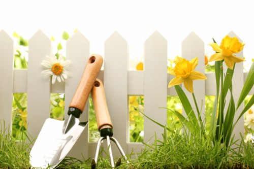 A close-up of a well-maintained garden border featuring vibrant yellow daffodils and white daisies growing alongside a white picket fence. In the foreground, there are gardening tools including a hand trowel and a small garden fork with wooden handles resting on the lush green grass. The background shows sunlight filtering through blurred greenery, indicating a bright, clear day suitable for outdoor gardening activities. The scene highlights typical elements of a suburban garden, with a neat lawn area and flowering plants that may be part of a professional gardening or lawn care service in Coney Hall or surrounding areas, such as Lawn Mowing Coney Hall specializing in landscaping and outdoor maintenance.